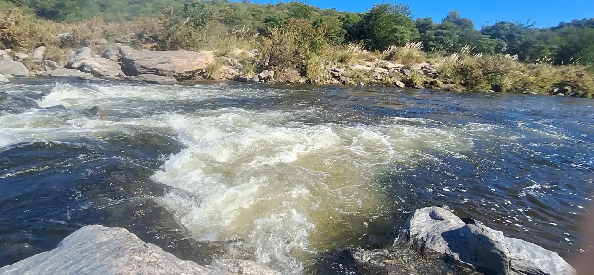 Rápidos del río Quillinzo entre rocas, con vegetación serrana en las orillas y cielo azul.